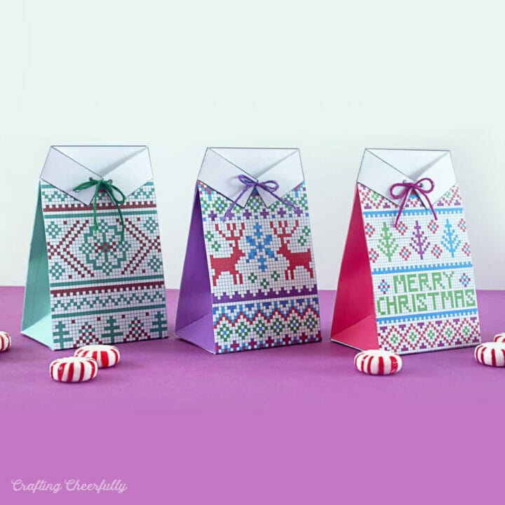 Colorful paper treat boxes designed to look like holiday sweaters sit on a pink surface.