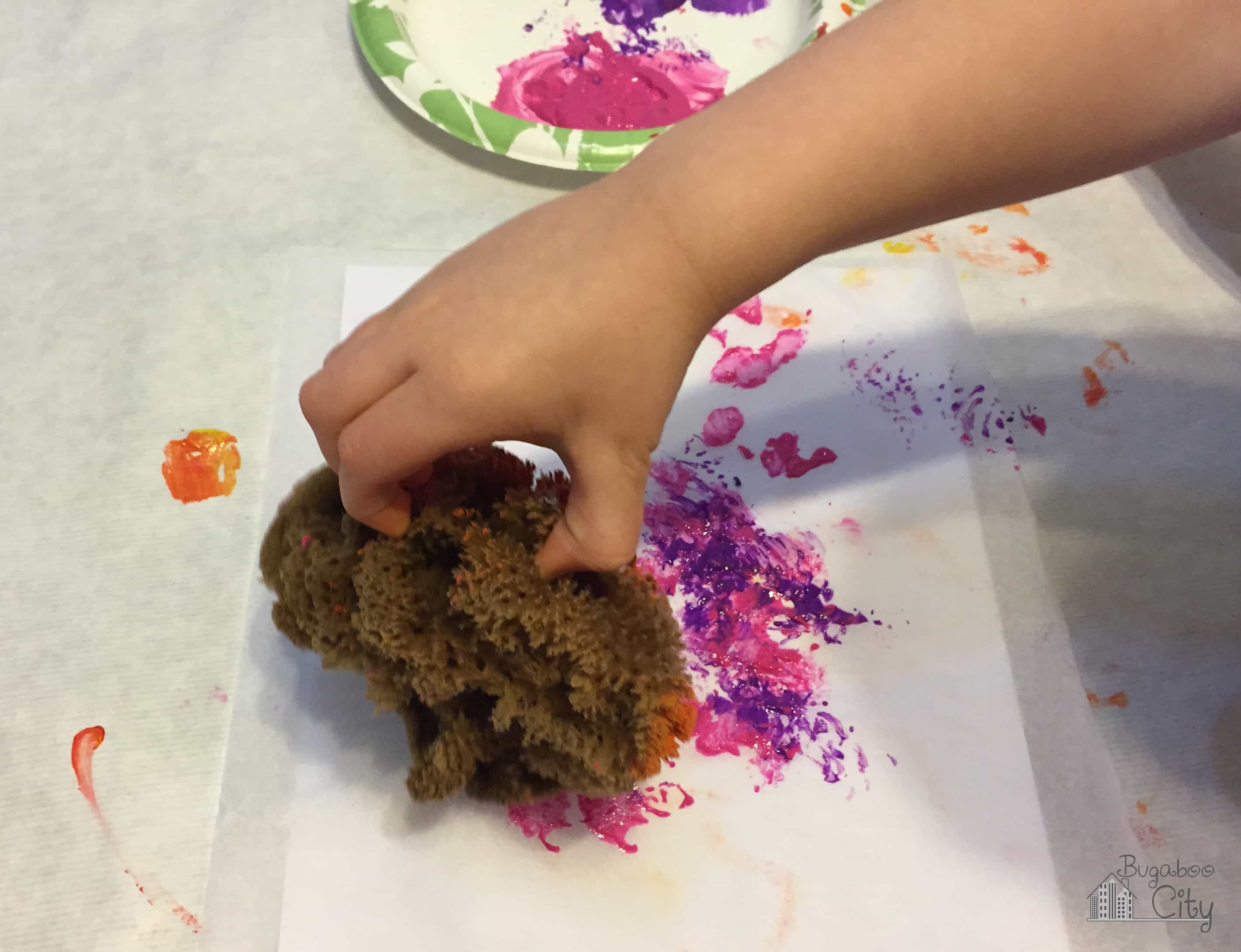 Child paints white tissue paper with pink and purple paint and a sponge.