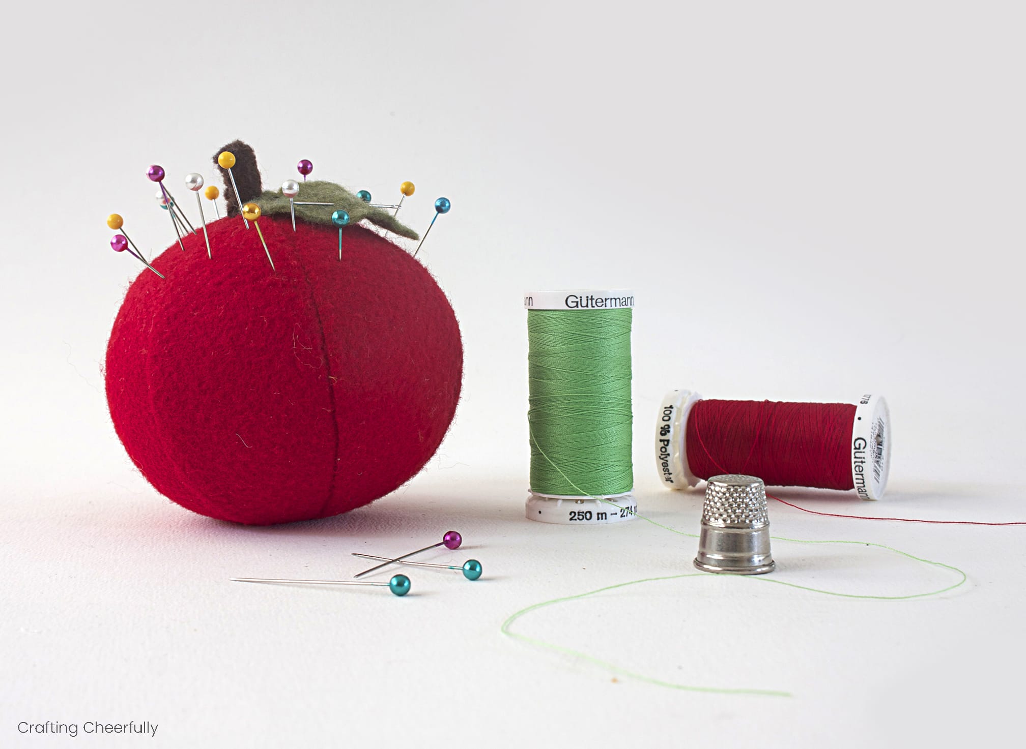 a red apple pincushion sits next to spools of red and green threads.