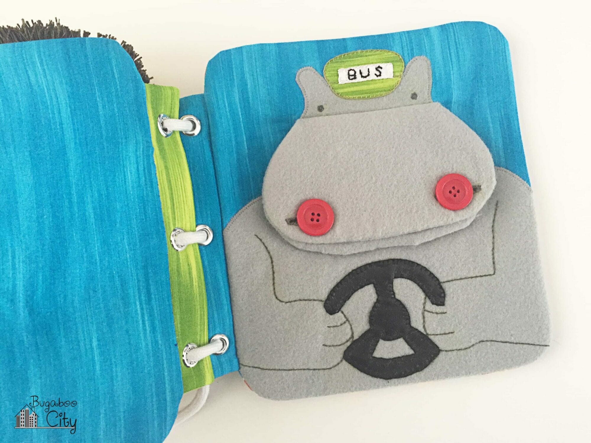 Fabric book page with a gray felt hippo wearing a hat that reads "BUS" and holding a pleather stealing wheel.