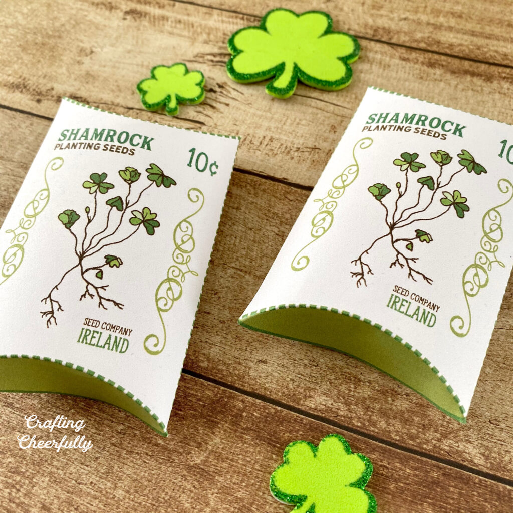 St. Patrick's Day pillow box treat boxes lay on a wooden table with little green shamrocks around them. The boxes have a picture of a shamrock plant on them with the words "Shamrock Planting Seeds" on them.
