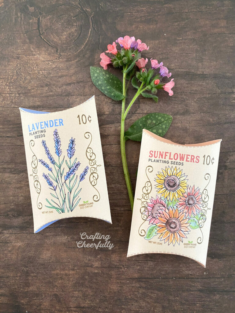Two sweet flower seed treat boxes laying on a wooden table with a pink flower next to them.