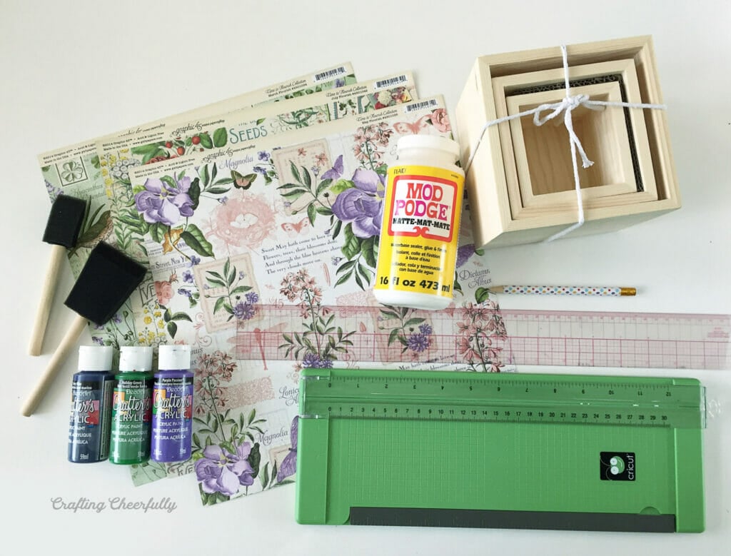 Supplies needed to make floral decoupage boxes are gathered on a white table including scrapbook paper, paint, a paper trimmer and wooden boxes.