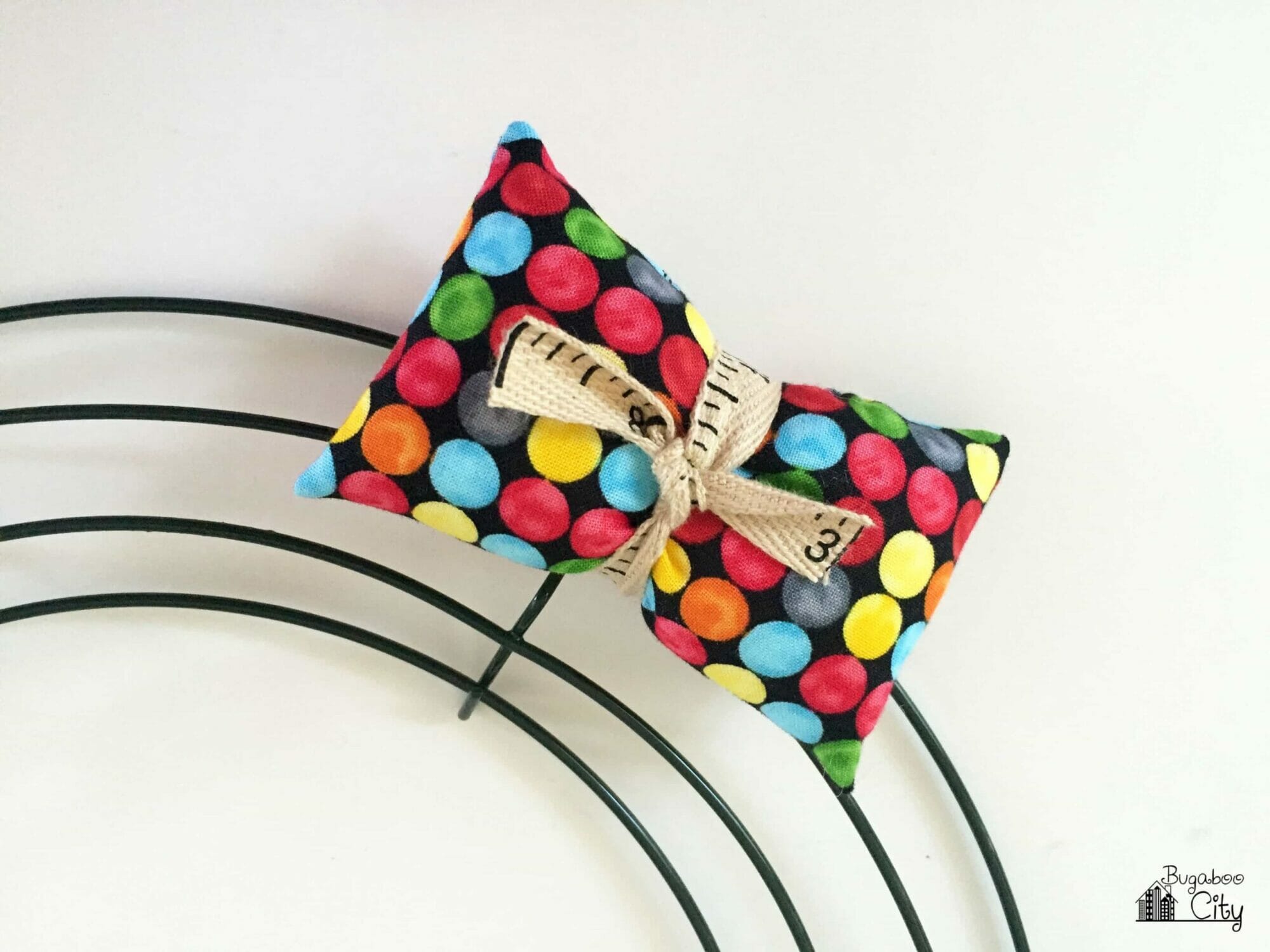 A multi-colored polk dot pillow is attached to the wire wreath frame with ruler ribbon.