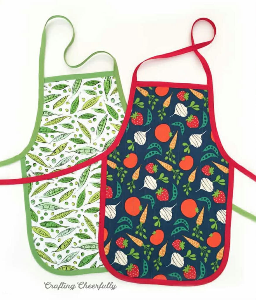 Two children's aprons with vegetable print fabric lay on a white table.