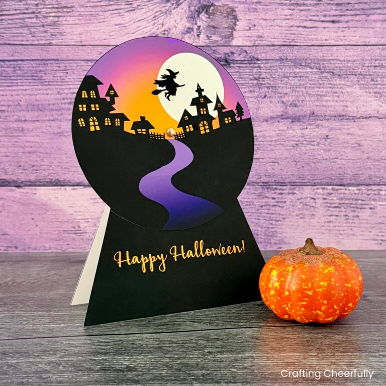 Halloween snow globe card sits on a black table with a small pumpkin.