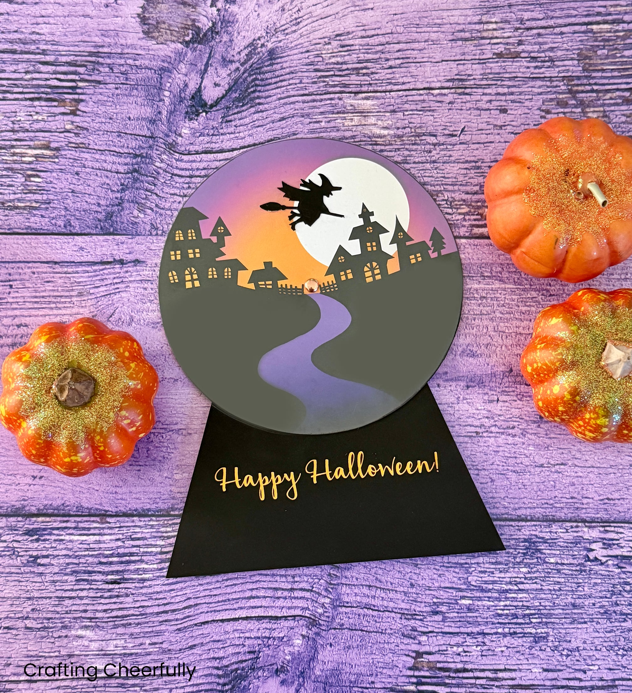 The Halloween snow globe card layson a purple wood surface with pumpkins around it.