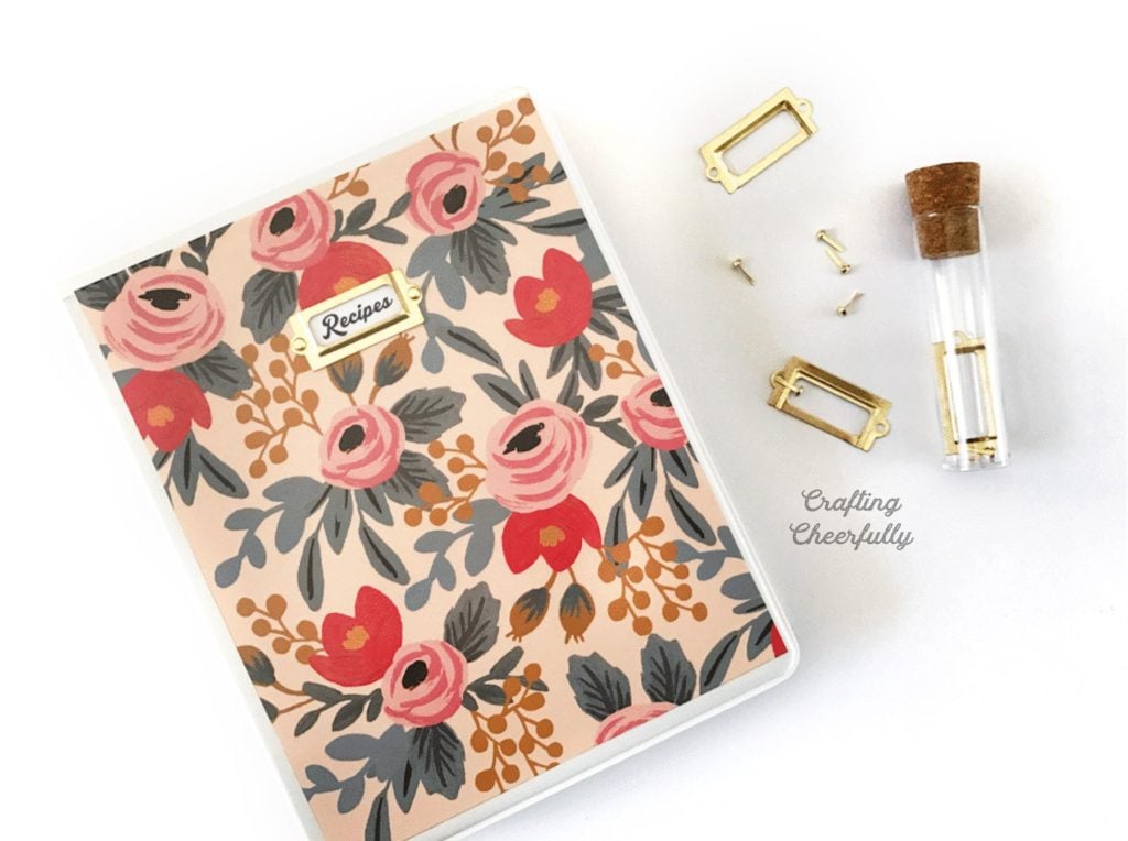 Small binder decorated with floral paper; a brass frame with the word recipe in it is on the front of the binder.
