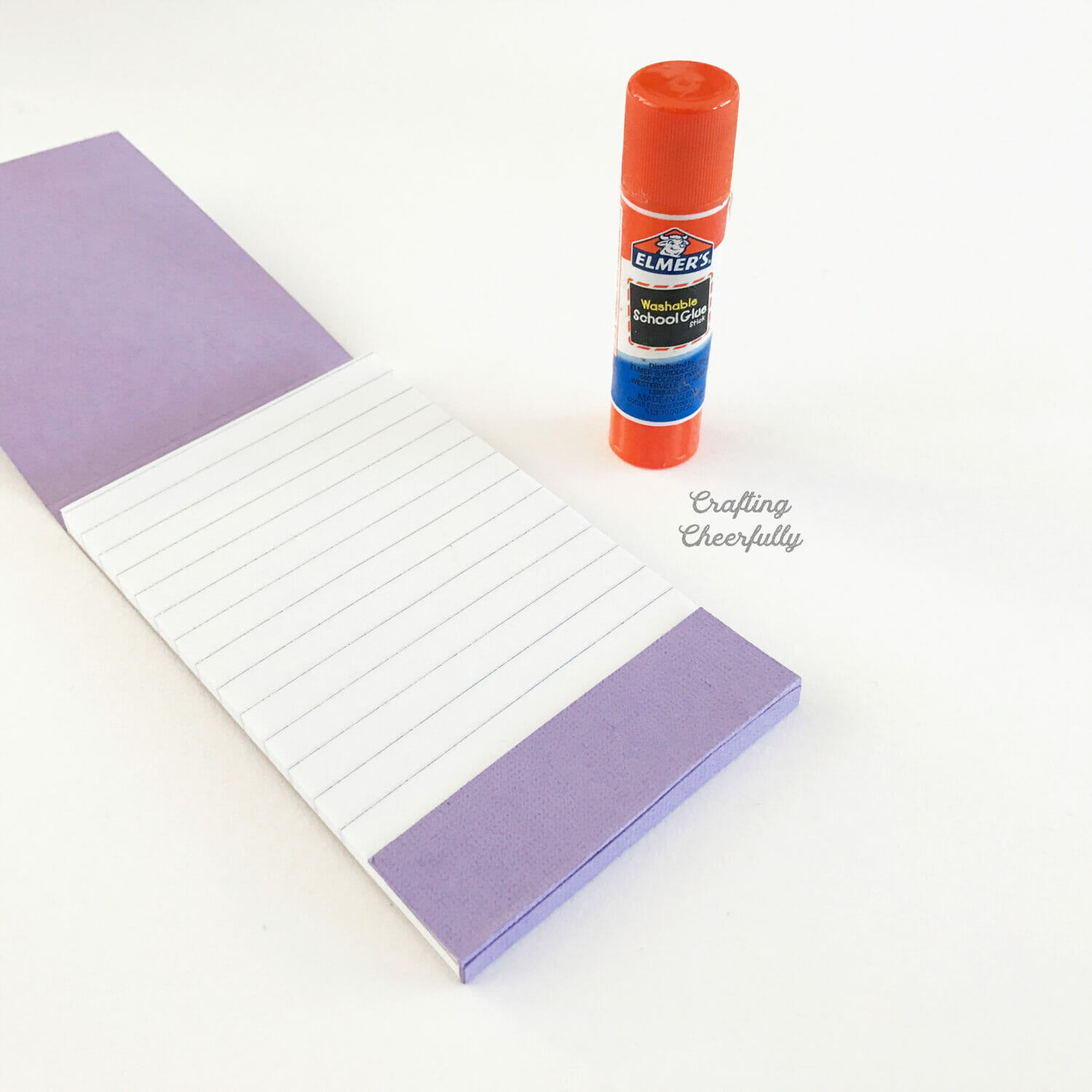 The outside of a notepad is wrapped in purple paper and sits next to a gluestick.