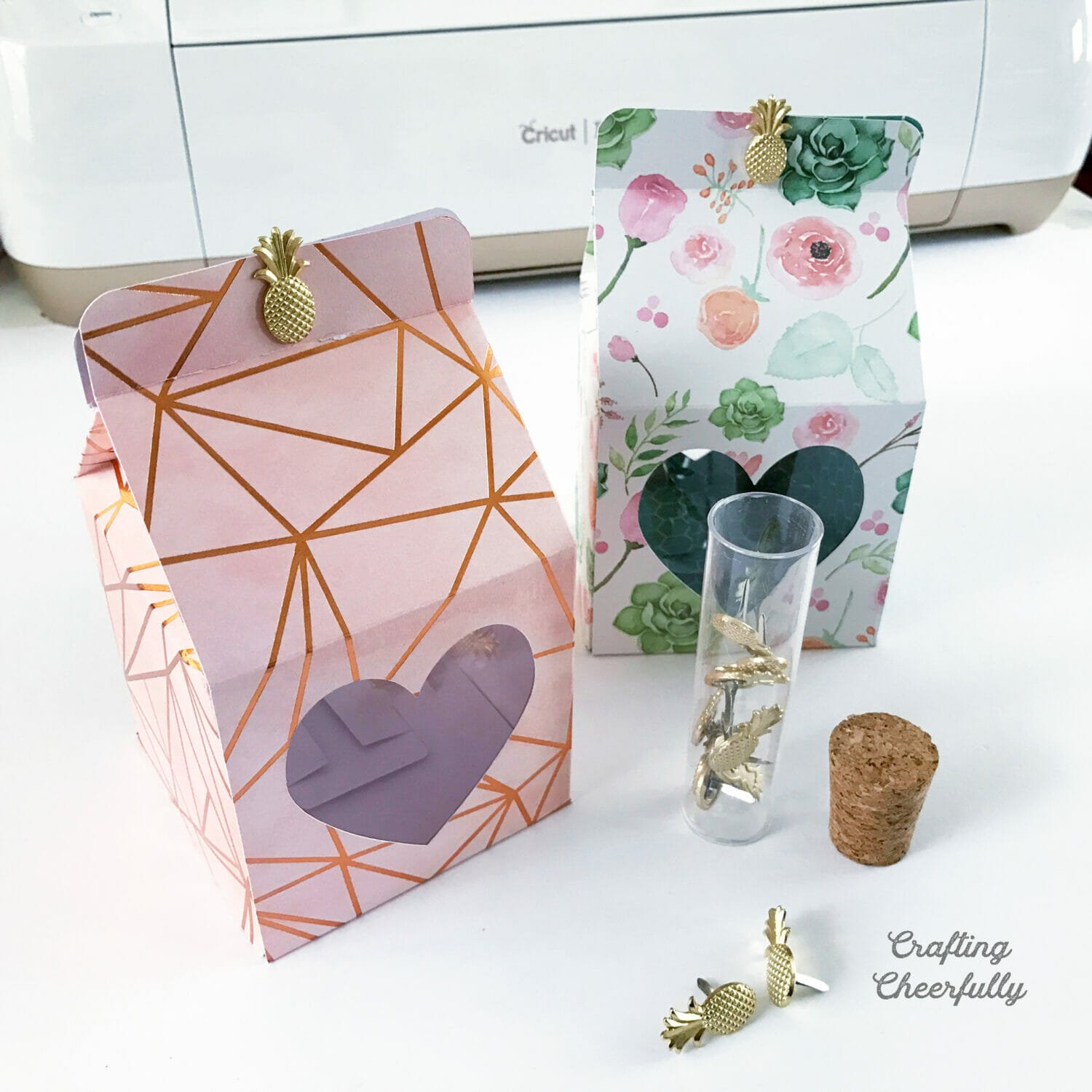 Two treat boxes with cut out heart windows sit in front of a Cricut cutting machine with a bottle of brads.