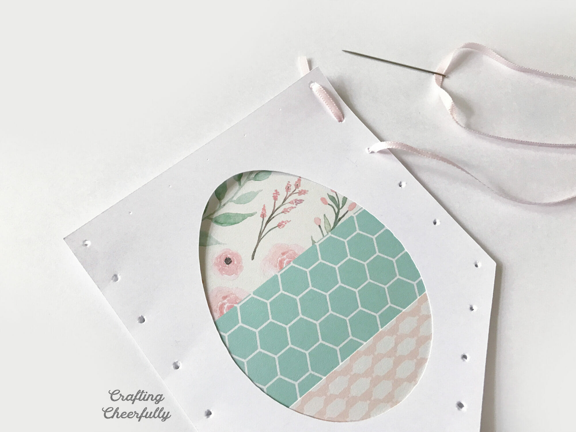A white pennant with decorative Easter egg is being stitched with thin pink ribbon starting from the top right corner.