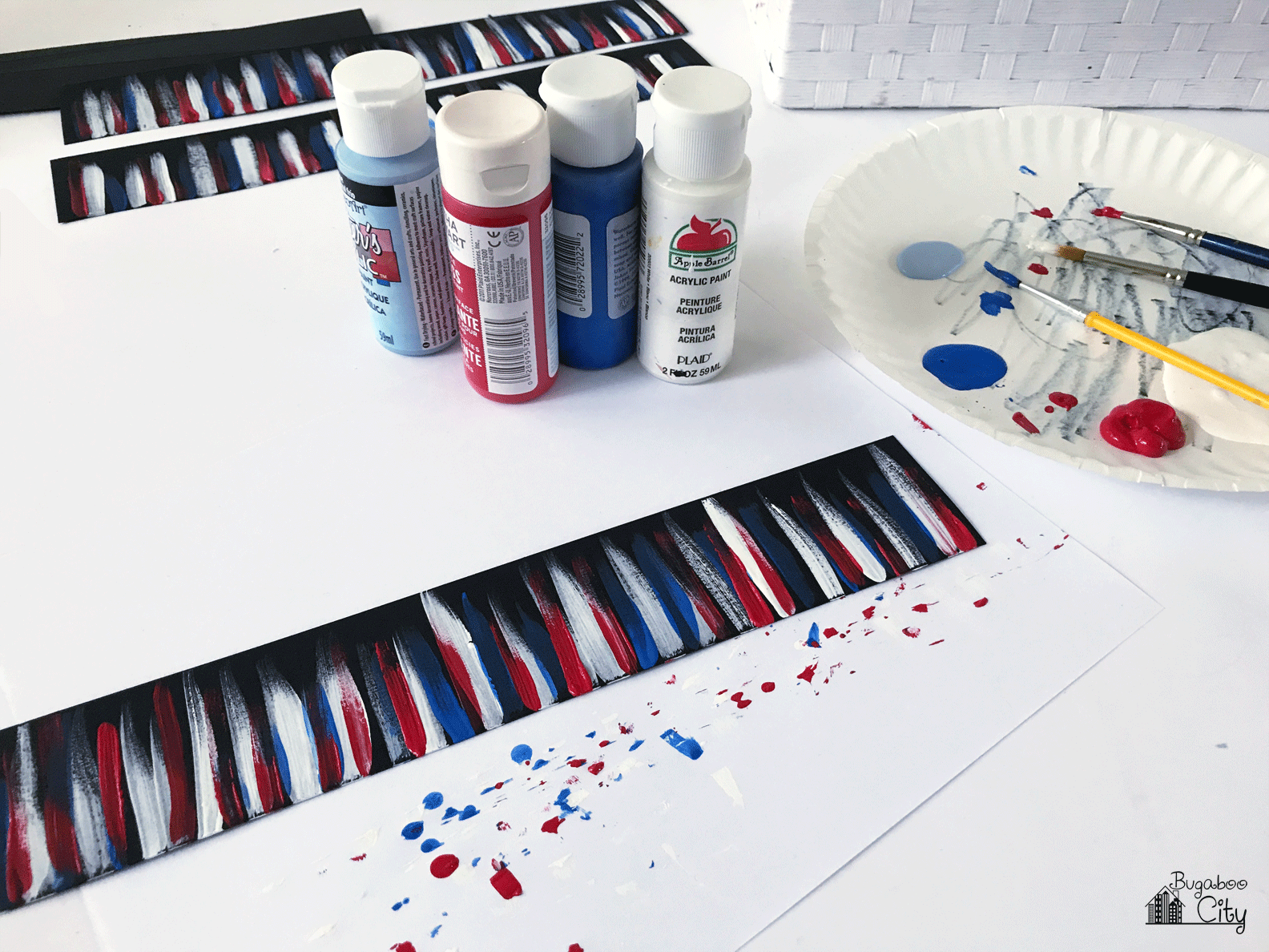 Bottles of acrylic paint sit next to a strip of black paper painted with streaks.