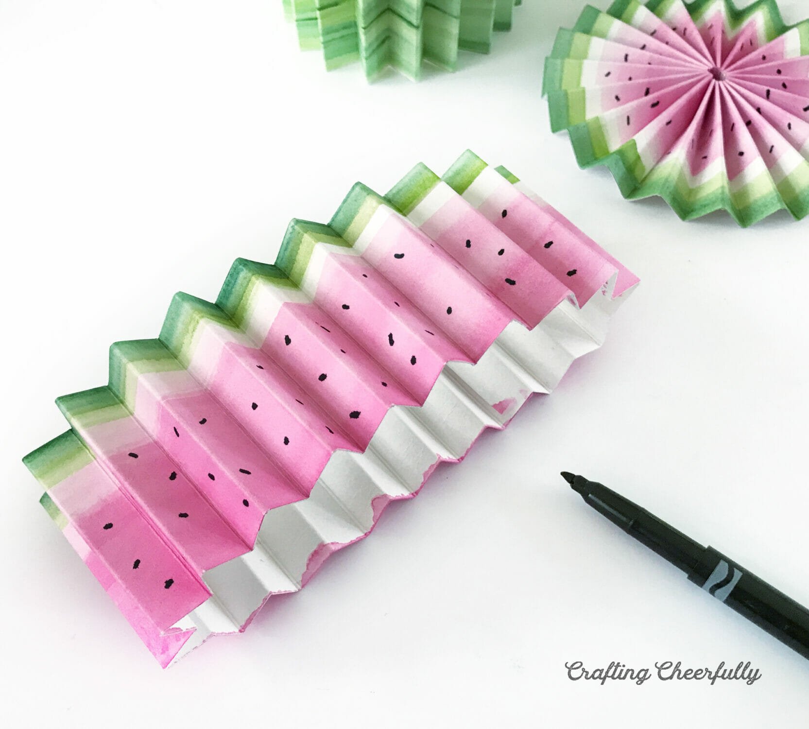 A black marker adds seeds to the pink area of the accordion-folded strip of paper.