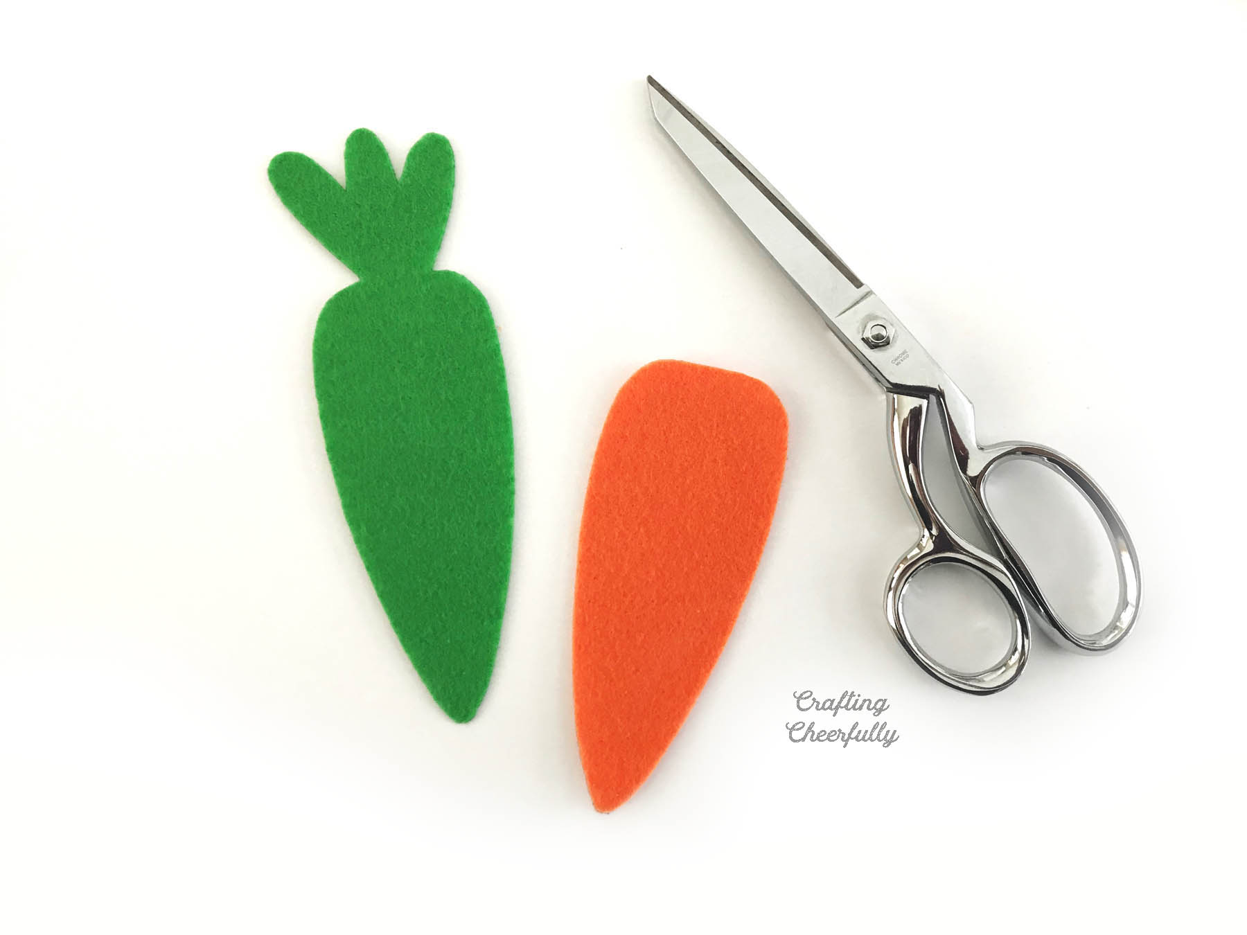 Green felt carrot with leaves lays next to an orange felt carrot and a scissors.