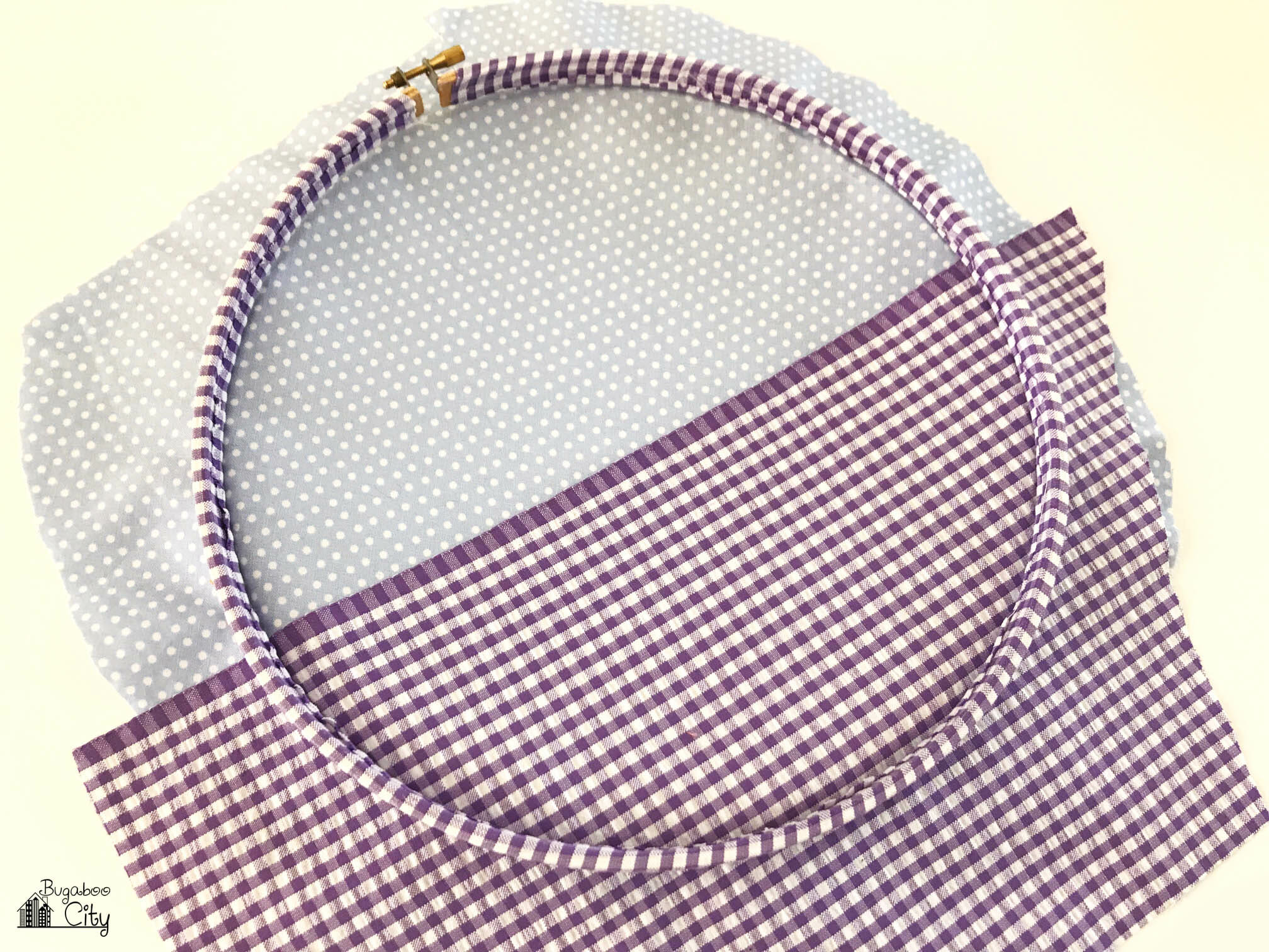 Embroidery hoop lays over two pieces of fabric; light blue on the top and purple gingham on the bottom. 