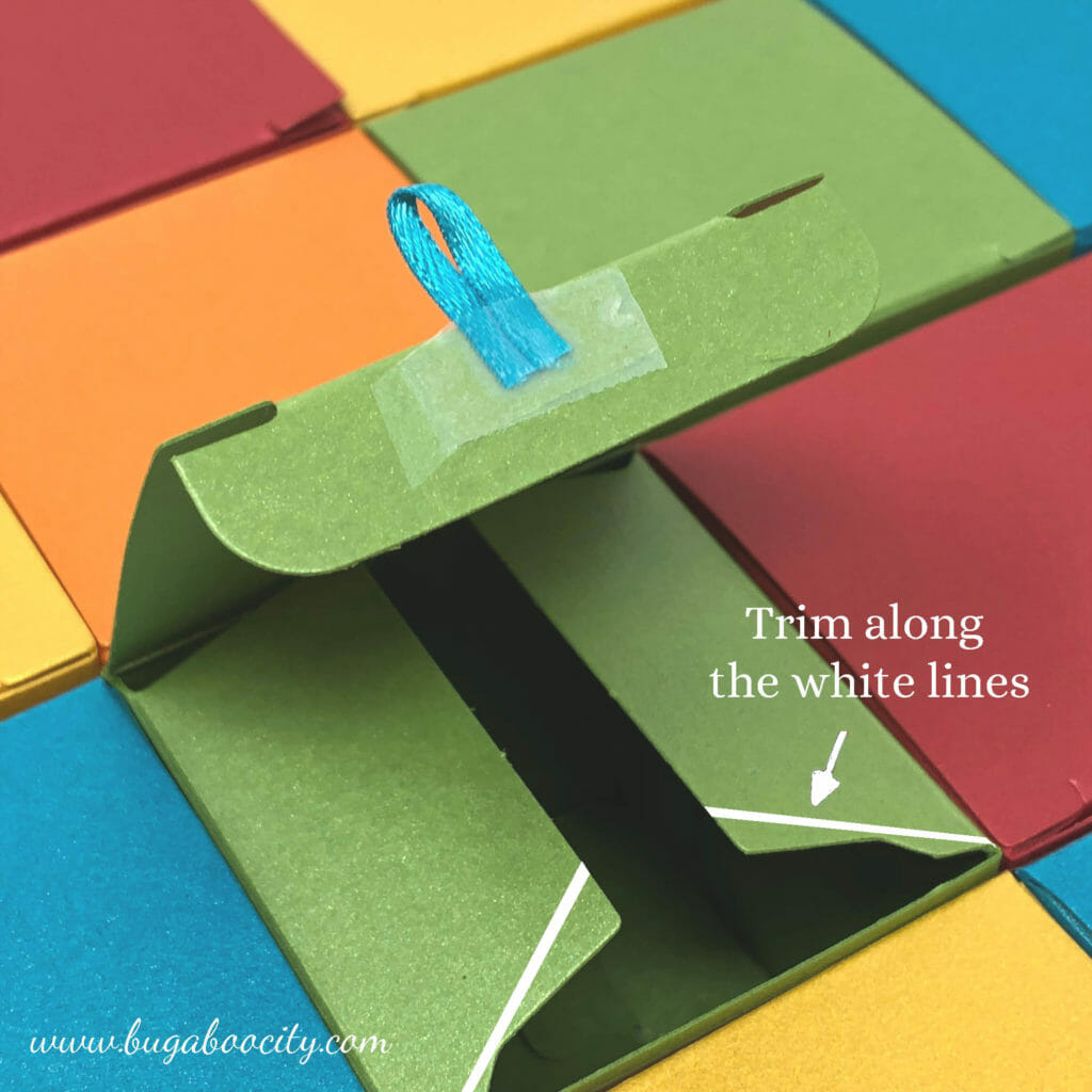 A ribbon loop is taped to the lid; white lines show where to trim the bottom of the box tabs.