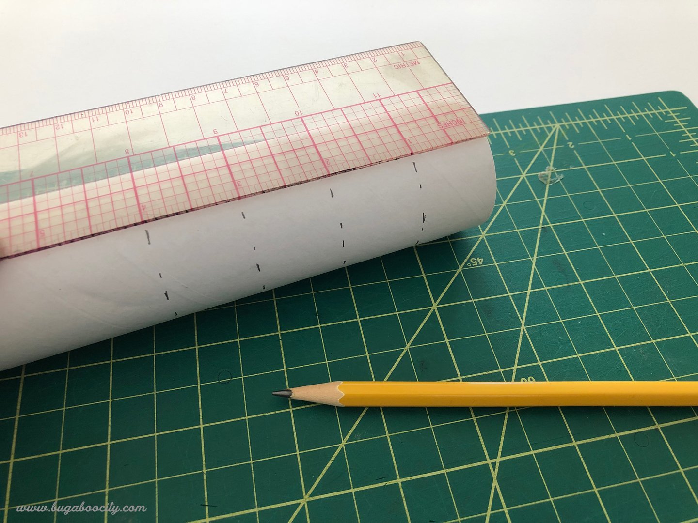 A clear ruler is laid over a white cardboard tube with notches marked on it in pencil.