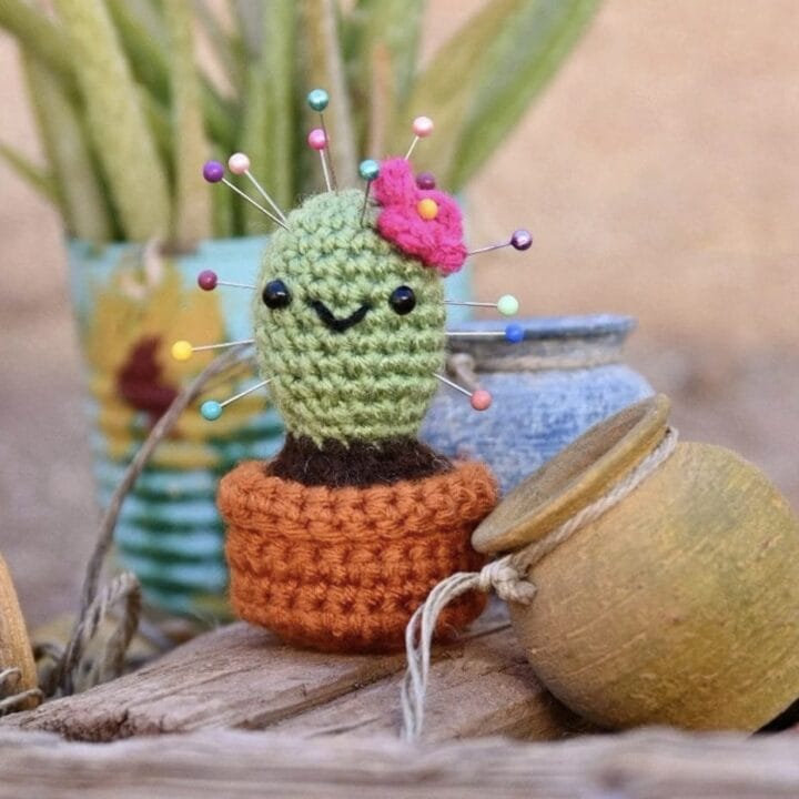Crochet cactus pin cushion with pin sticking out of it sits near a real cactus.