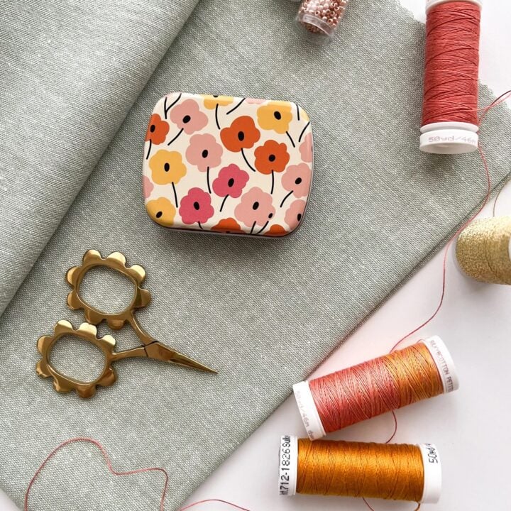 Floral magnetic needle tin rests on fabric next to thread and a mini scissors.