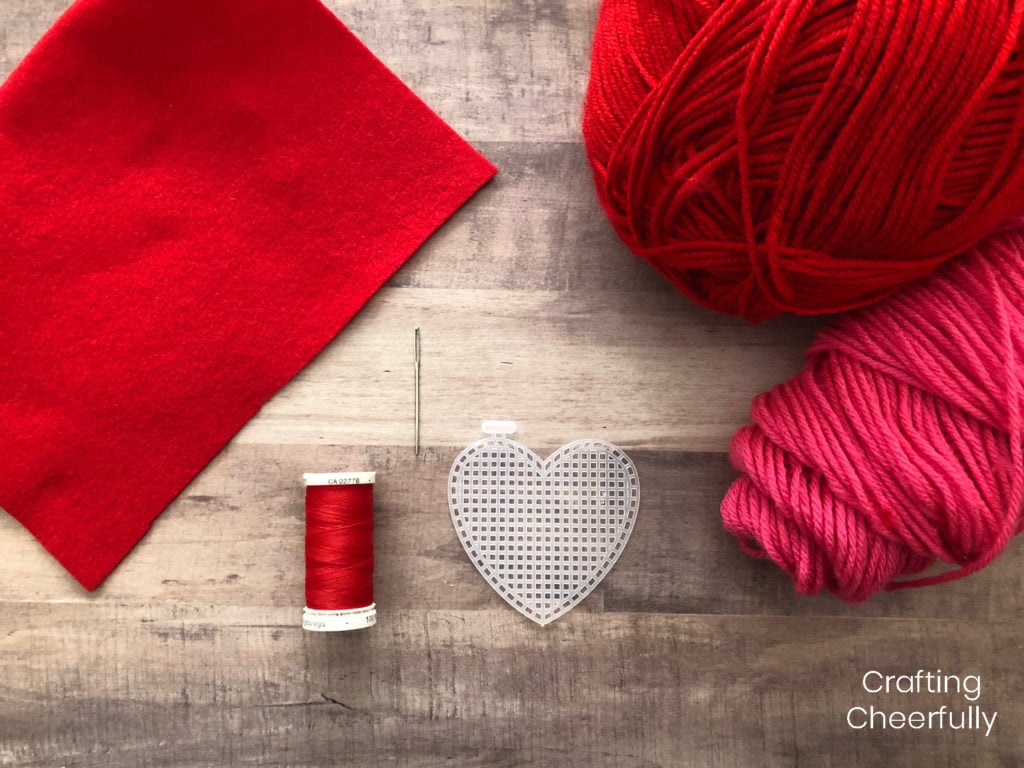 Supplies including red felt, red thread, needle, red and pink yarn and a heart-shaped plastic canvas. 