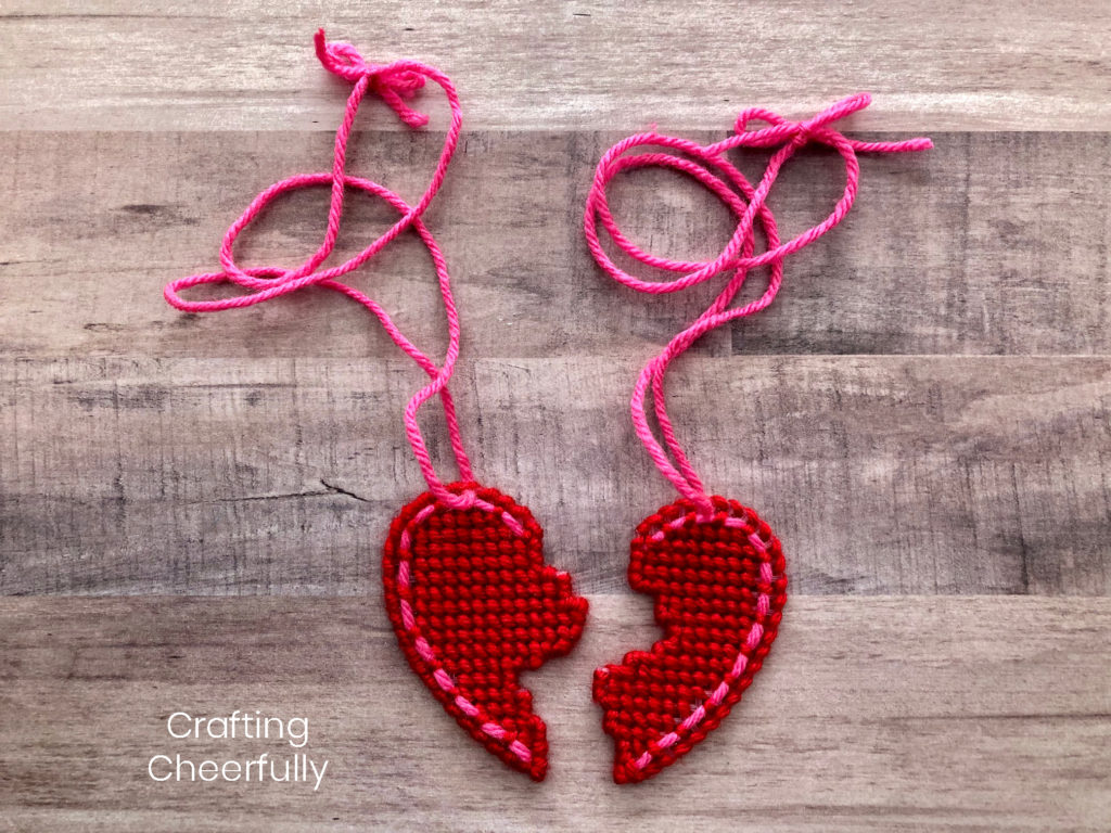 Two halves of a heart made from plastic canvas and stitched with yarn lay next to each other; each has a pink yarn necklace attached to the top.