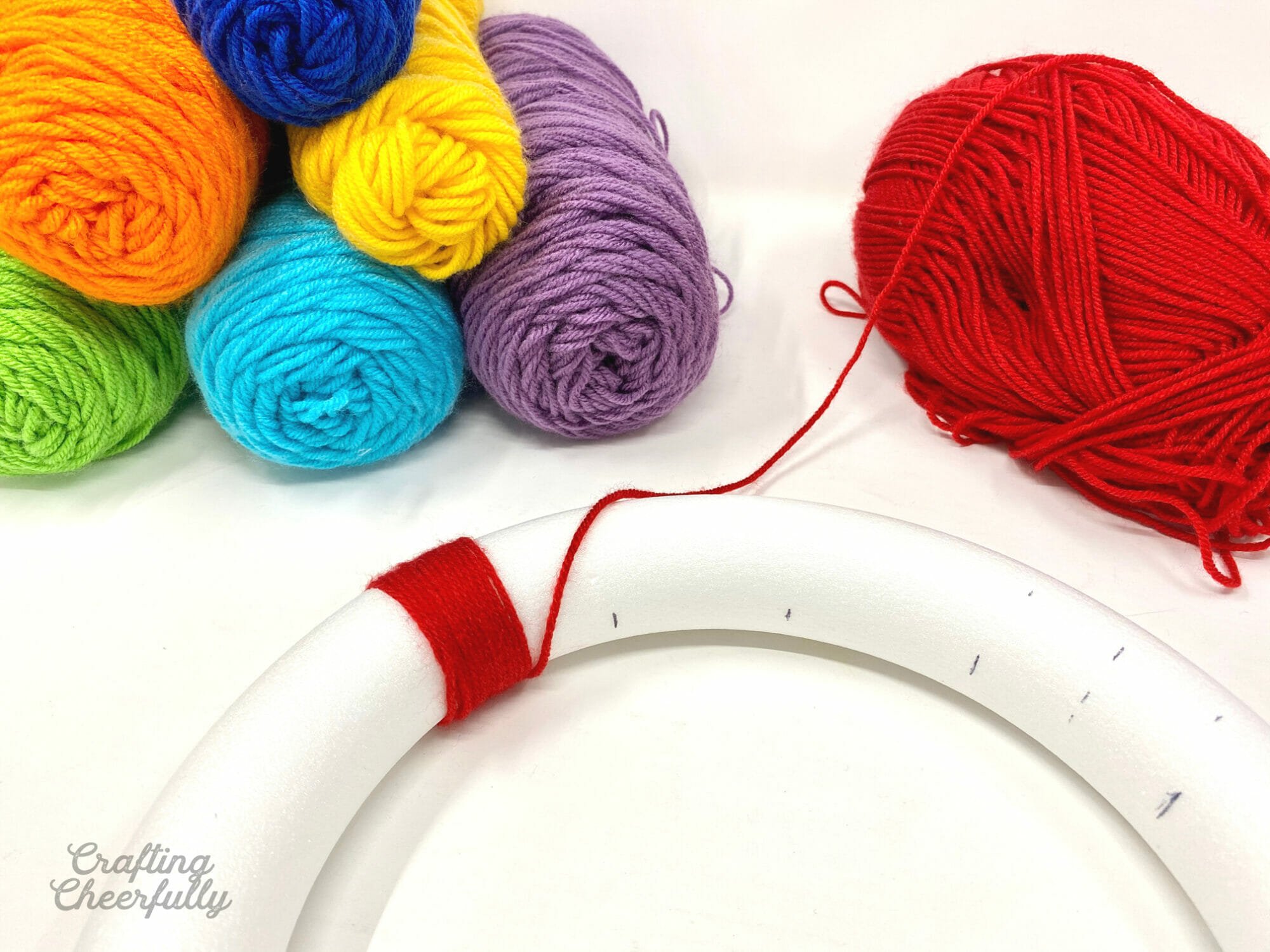 Red yarn is starting to be wrapped around the wreath form; rolls of colorful yarn sit nearby.