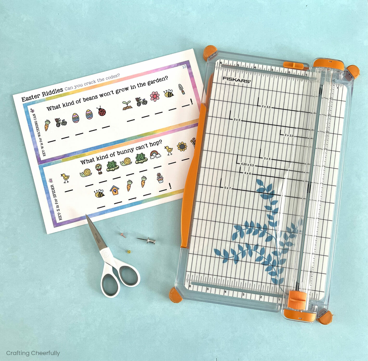 Printable riddle worksheet lays next to a paper trimmer, scissors, push pin and brads.