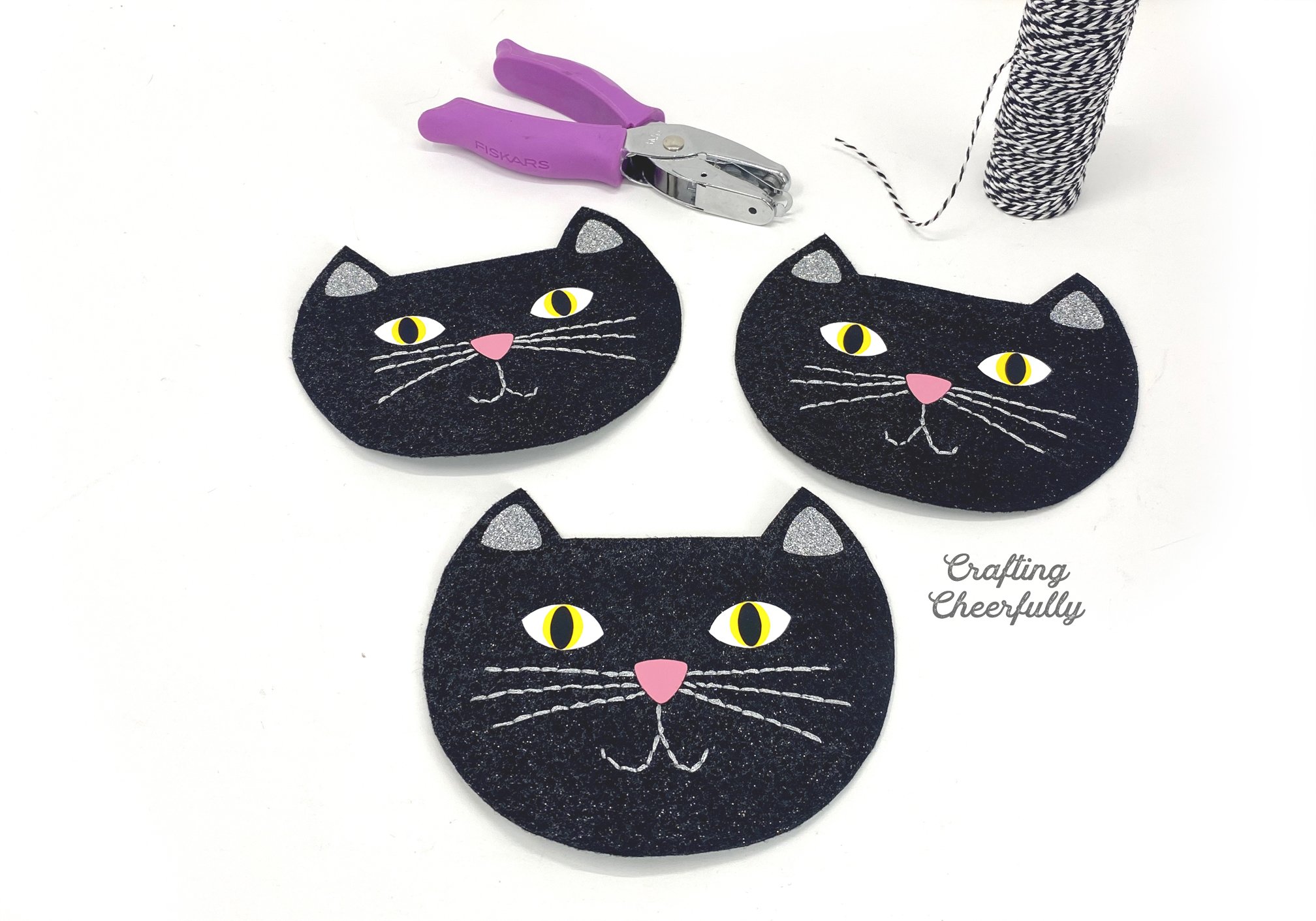 A hole punch and a roll of black and white Baker's Twine lays next to three black cat felt shapes.