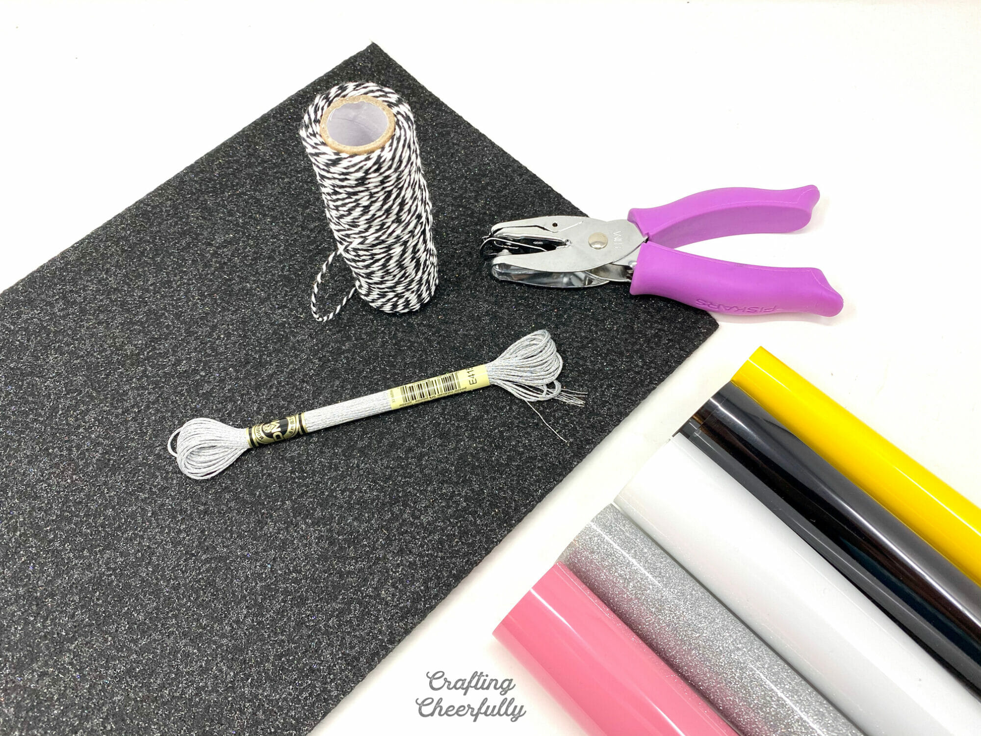 Rolls of Iron On in pink, silver, white, black and yellow lay next to a sheet of black glitter felt, a whole punch, Baker's Twine and silver embroidery floss. 
