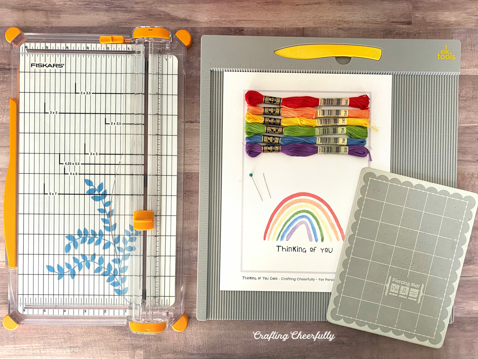 Supplies and tools including a paper trimmer, scoring board, piercing mat, embroidery floss and printable rainbow card.