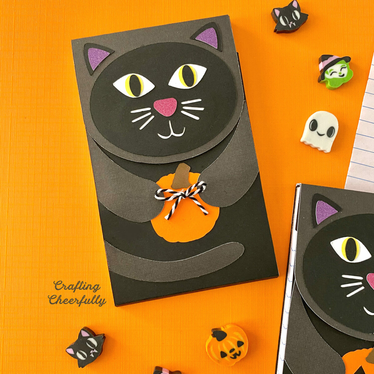 A black cat paper notepad holds a small orange paper pumpkin with a tiny bow out of twine on it.