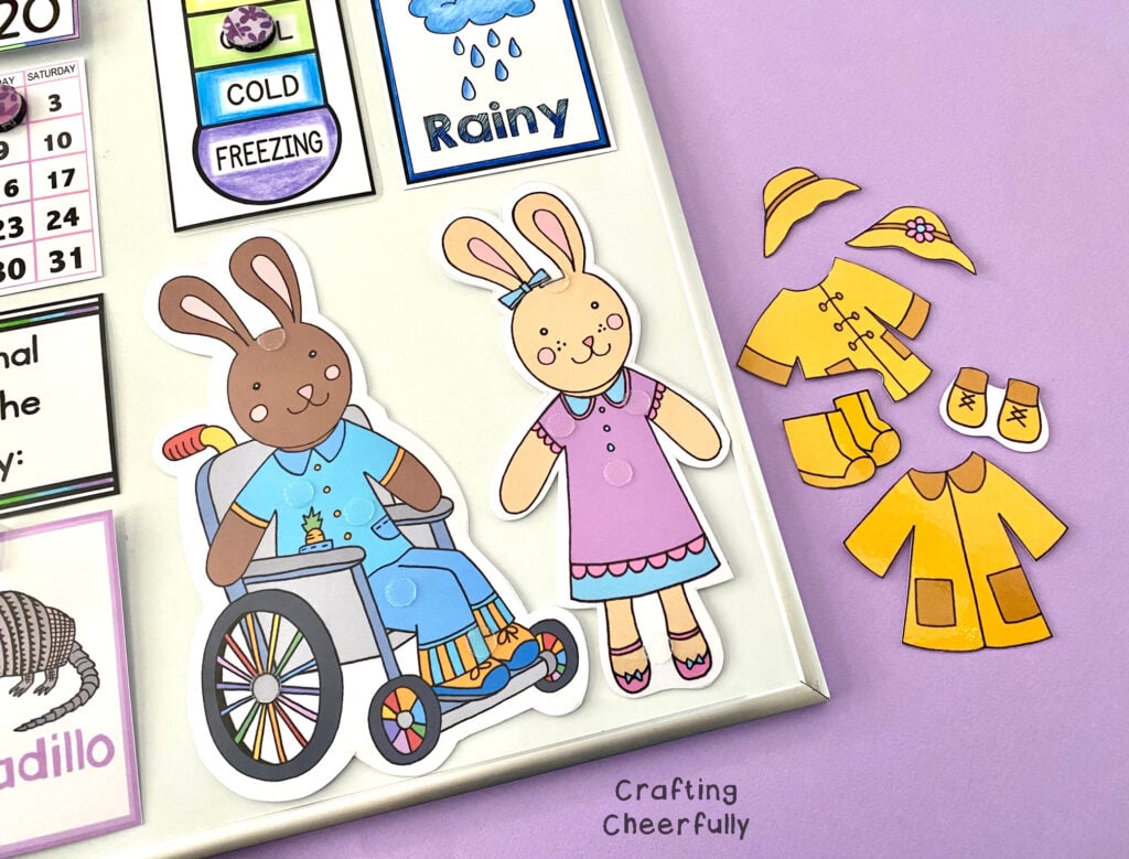 Two bunny dolls; one with brown fur using a wheel chair, and the other with cream fur. There are yellow rain clothes laying next to the board.