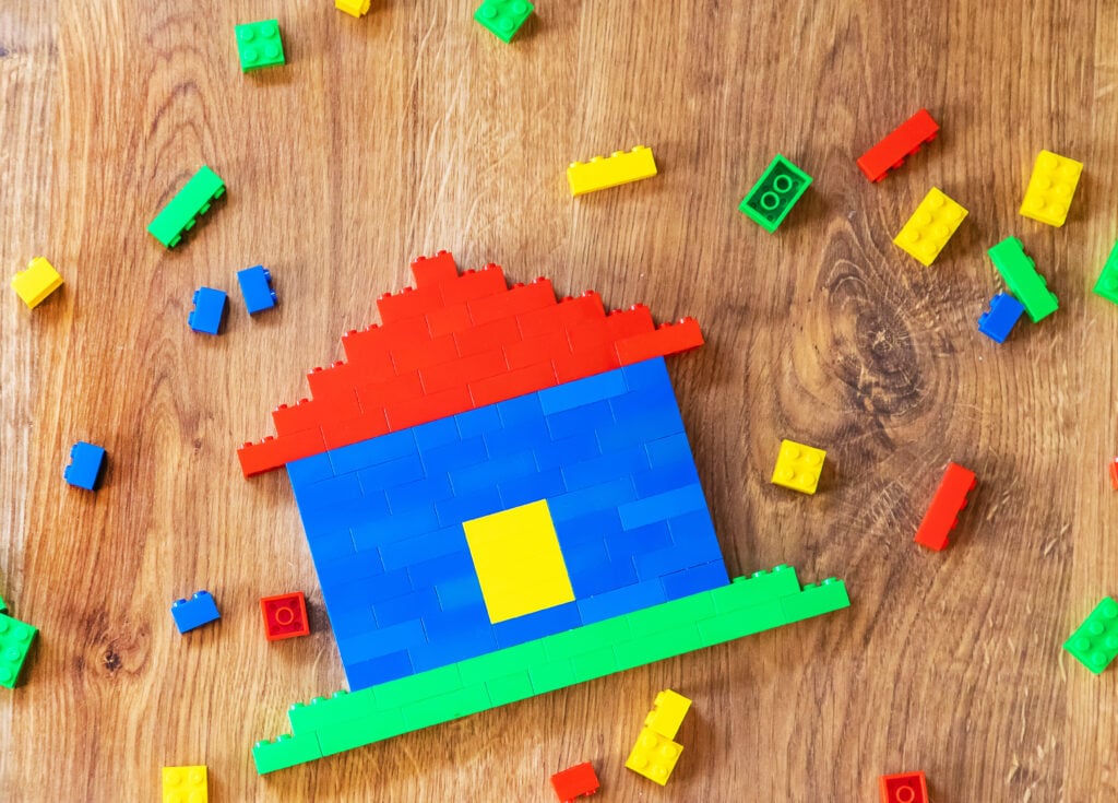 A two dimensional house built from LEGO lays on the floor surrounded by LEGO pieces.