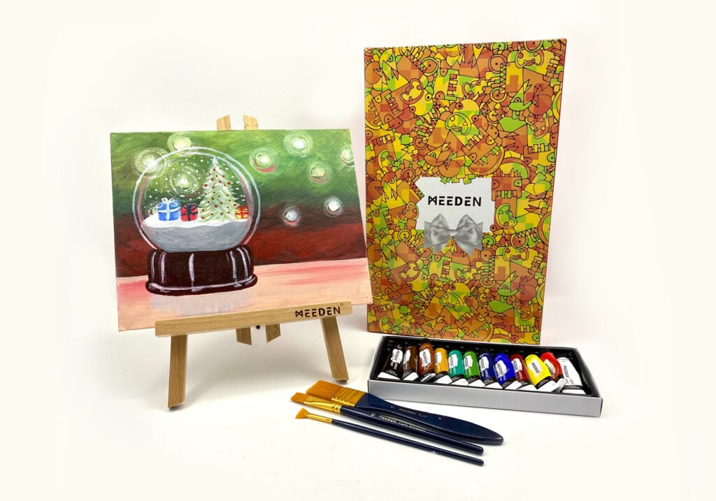 A painted canvas with a holiday snowglobe on it sits on a mini easel next to acrylic paints.
