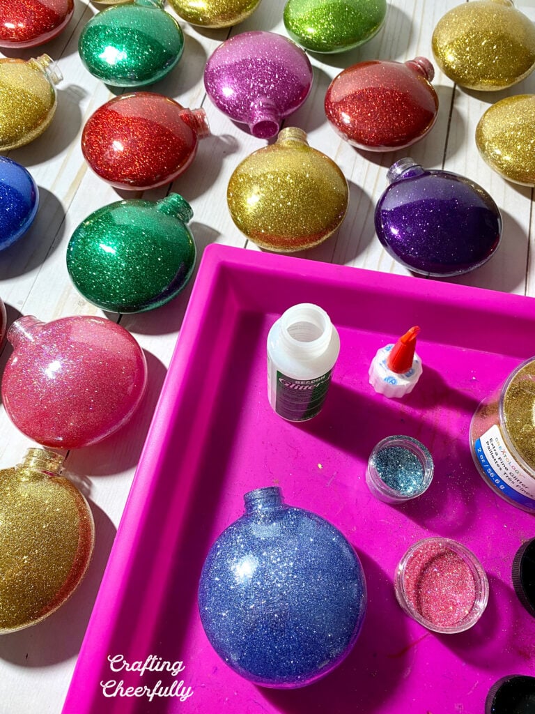 Glitter ornaments of all different colors lay on a table after being filled with Glitter-It adhesive and glitter. A pink tray holds containers of glitter.