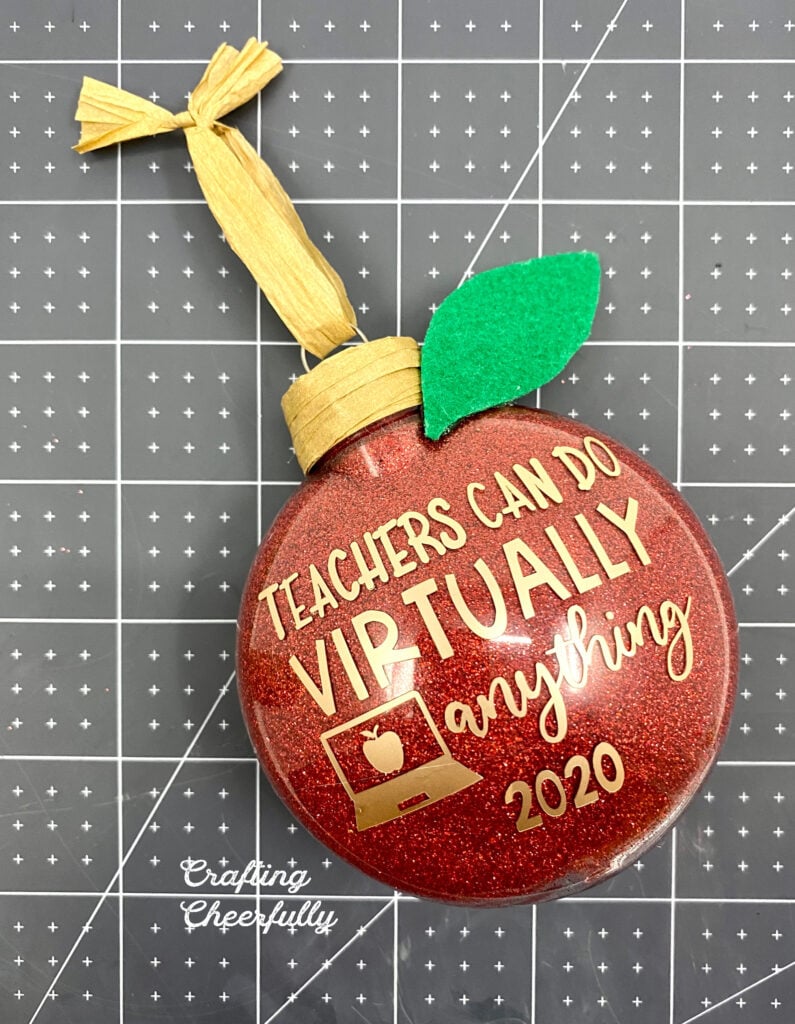 Red glitter ornament with brown raffia wrapped around the top of the ornament. A green felt leave at the top of it. Brown raffia hanging loop.