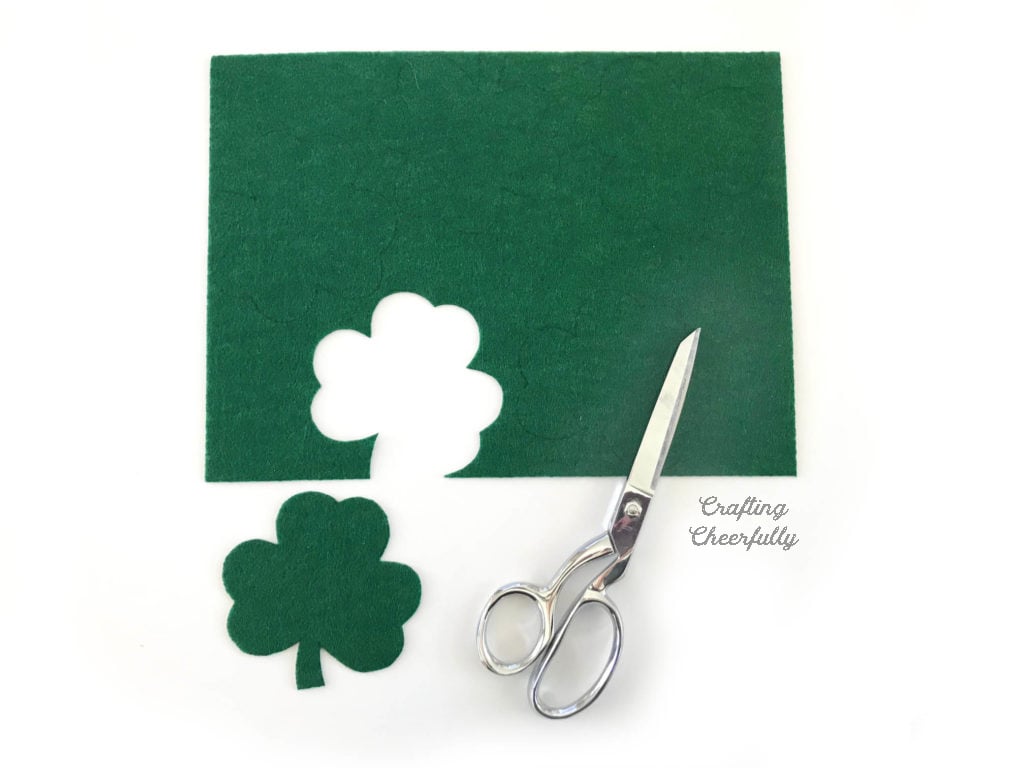 Green felt with a shamrock cut out. Gingher sewing scissors lays near the felt.