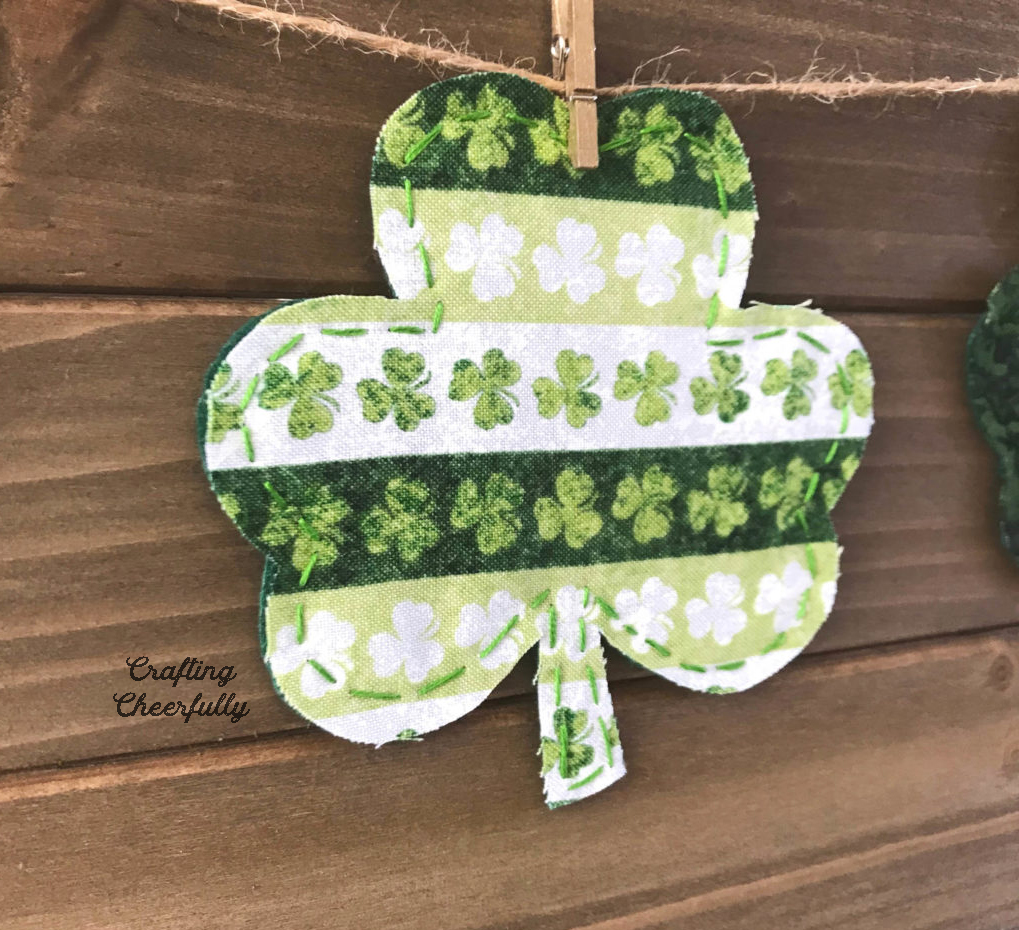 A fabric shamrock that is handstitched around the edges hangs on a board with a clothespin.