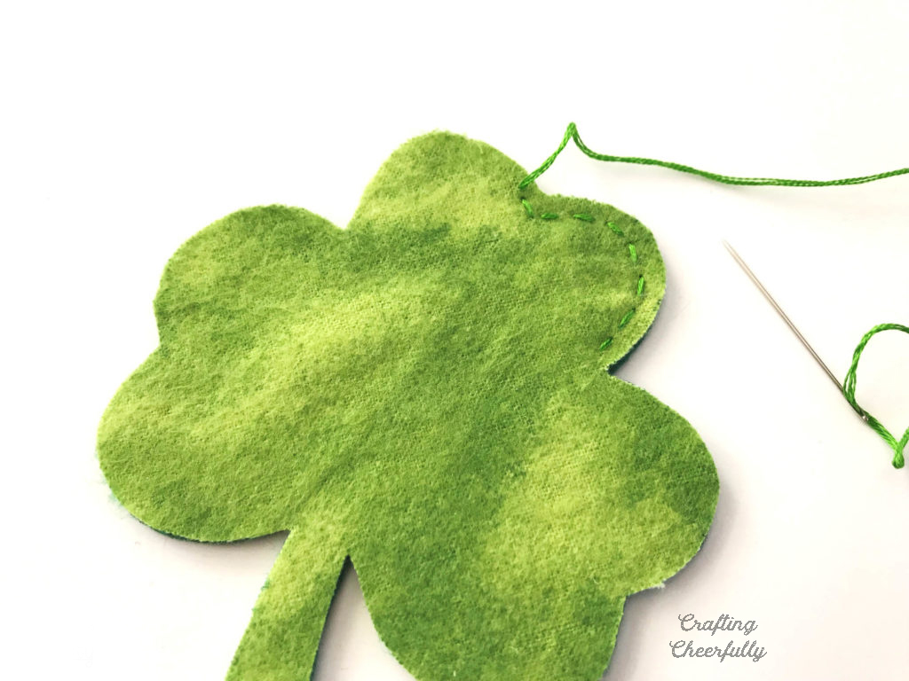 A fabric shamrock is being stitched around the edges with a needle and green embroidery floss.