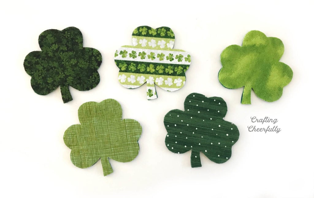 Five fabric shamrocks lay on a white table. The fabric layer is on top of the felt layer.