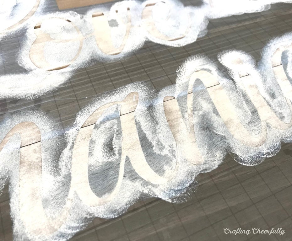White paint is applied lightly and unevenly to a stencil on a wooden board.