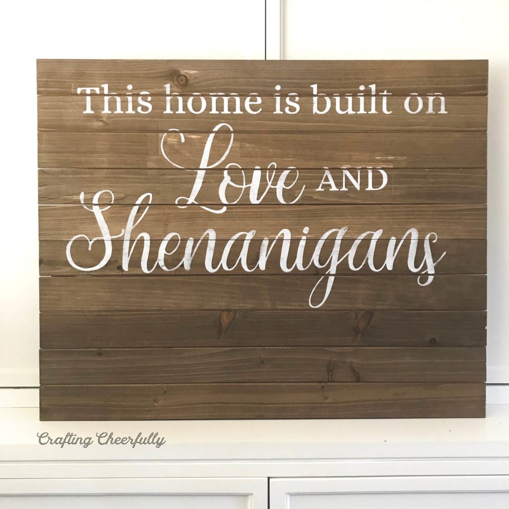 A wooden sign with white words painted on it leans against a white cupboard.