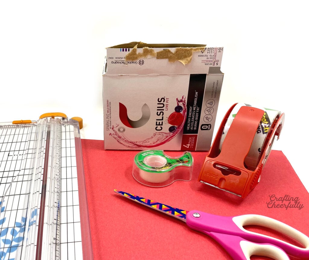 A cardboard box, scissors, tape and red construction paper sit on a table ready to craft with!