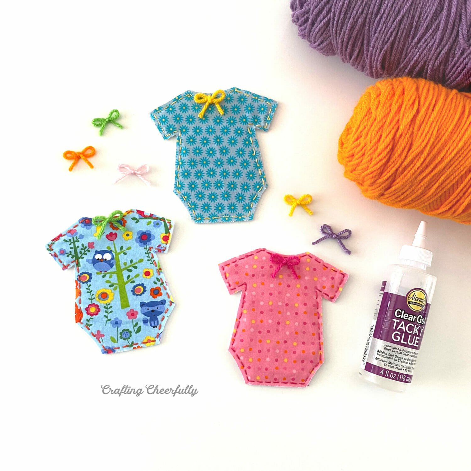 Cute fabric onesies for the banner lay next to yarn, tiny yarn bows and glue.