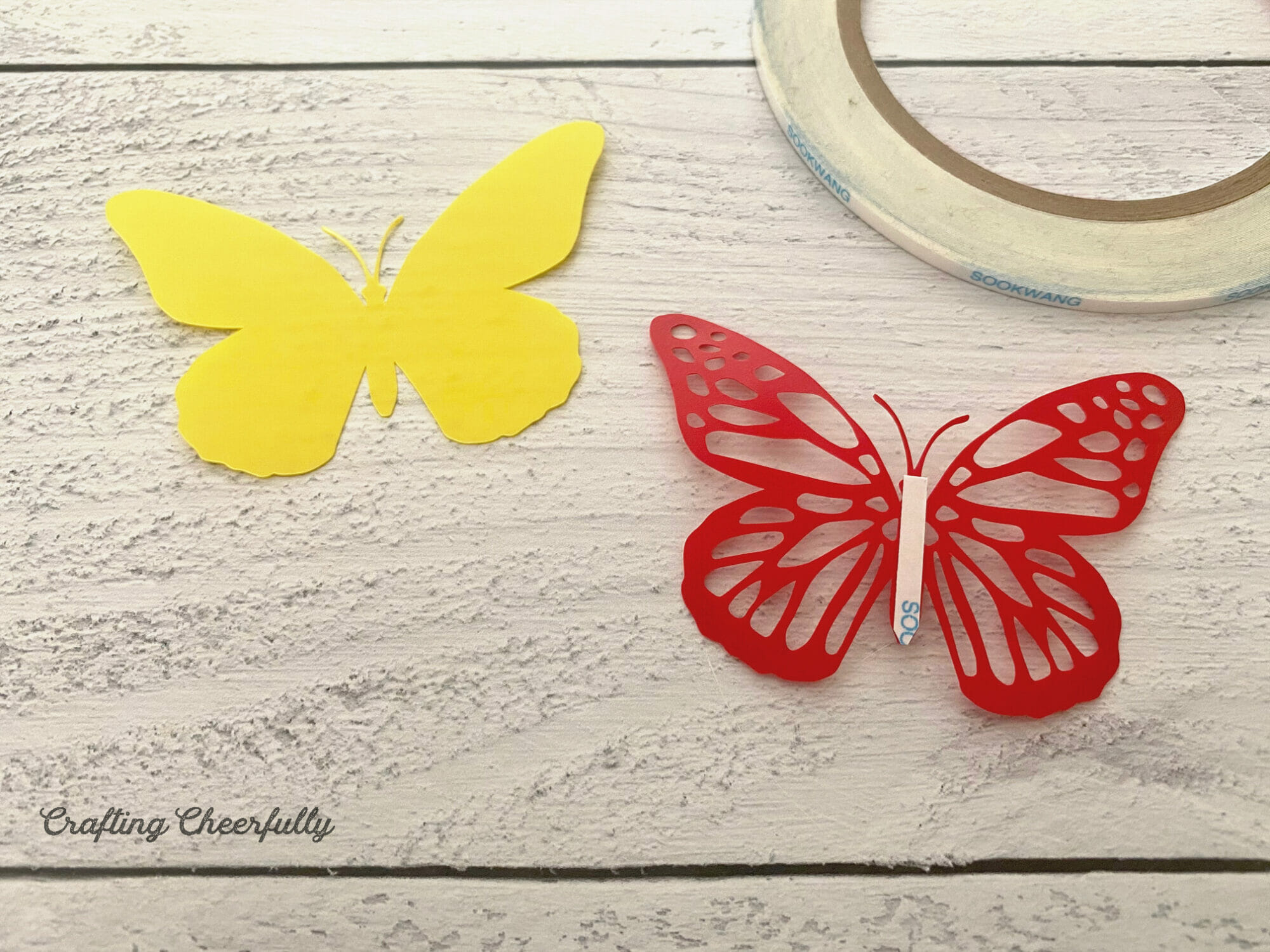 A yellow butterfly and a red butterfly with a cut out design lay next to each other, double-sided tape is on the back of the red one.