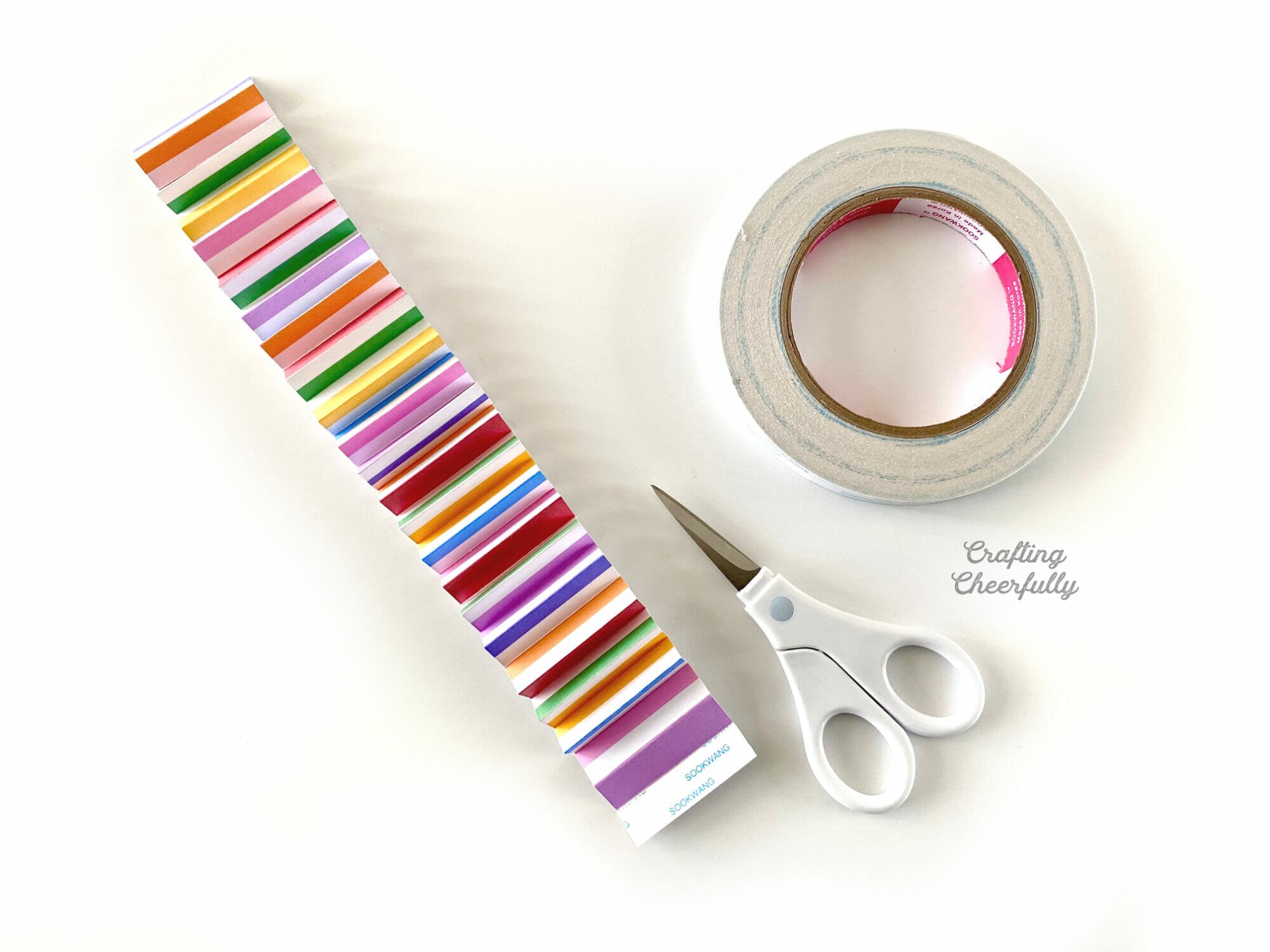 Strip of colorfully striped paper accordion folded with a smalls scissors and a roll of double-sided tape laying next to it.