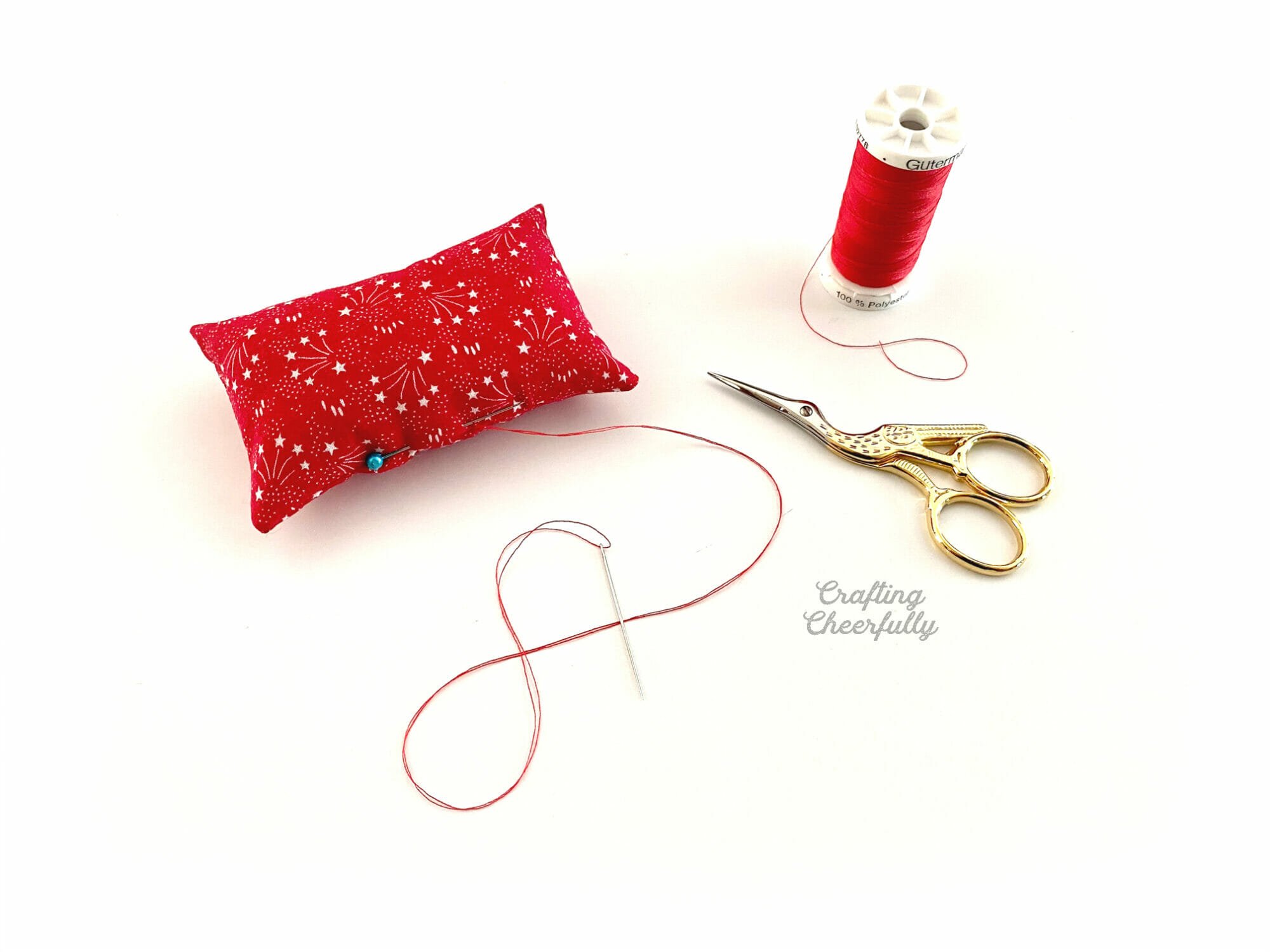 Red pillow with a pink in it laying next to a red spool of thread and stork scissors.