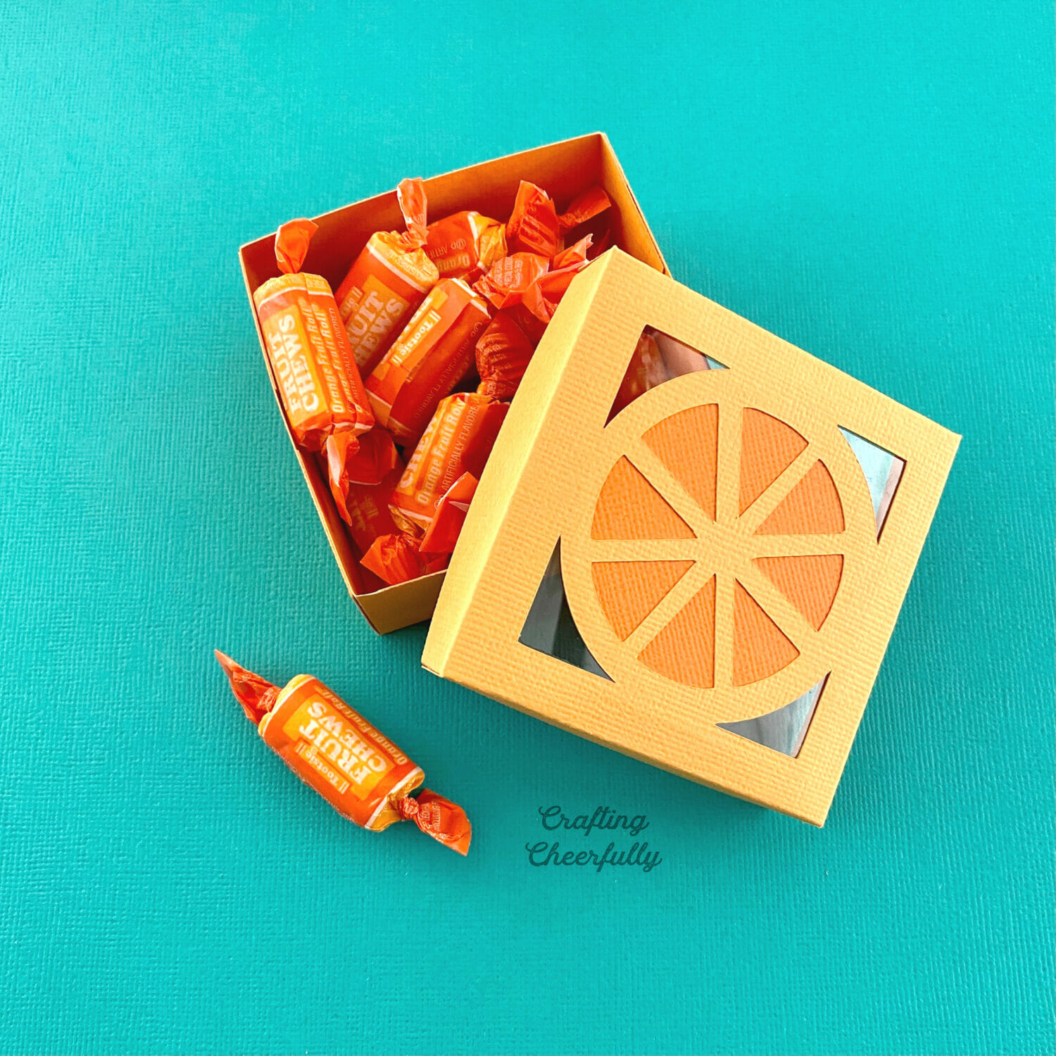 Orange tutti frutti box filled with orange candy on a turquoise background.