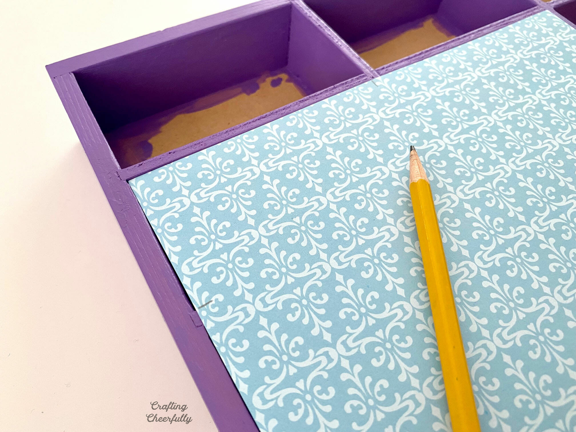 A sheet of blue patterned paper is laid on top of the purple display case and the width of a cubby is marked with pencil.