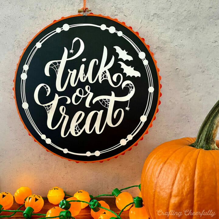 Halloween embroidery hoop hangs on a wall with a pumpkin and pumpkin lights underneath it on a tabletop.